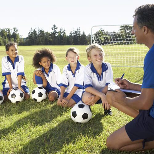 Male with clipboard and four young children holding soccer balls.